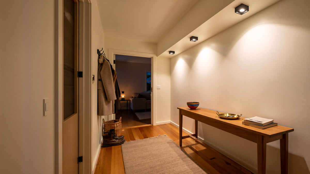 Warm downlights casting soft pools of light on a plaster hallway wall above an oak console, with a lamp glowing beyond